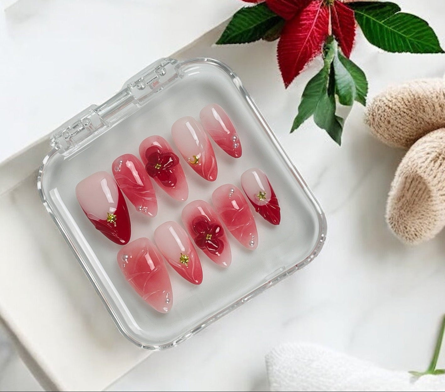 A set of red and pink press-on nails with gold glitter accents, displayed in a clear plastic case on a white marble surface with a red poinsettia and beige knit accessories.